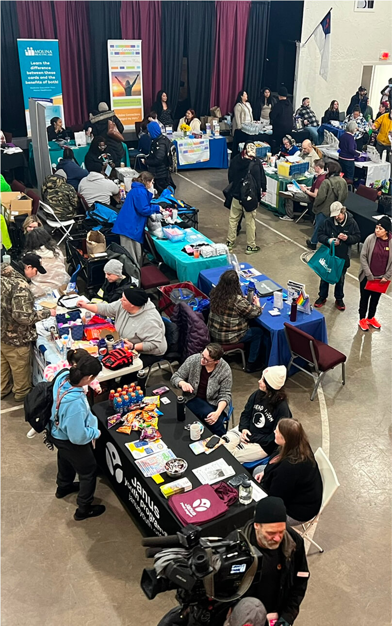 An overhead photo shows the 20+ community partners who are tabling a Project Homeless Connect event, which is held annually. This event is for people experiencing homelessness, and offers free health care services, help with filling out housing request forms, pet care, and provides food and clothing and supplies.